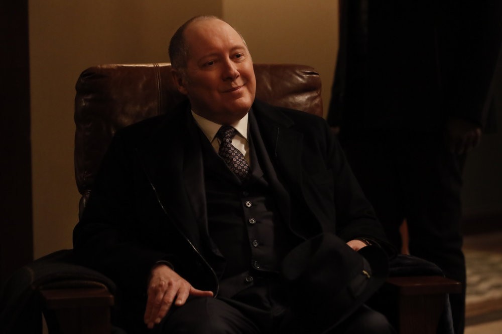 Raymond Reddington (James Spader), un sourire satisfait sur les lèvres, est assis dans un fauteuil