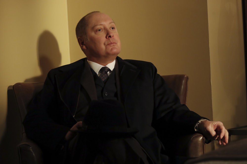 Raymond Reddington (James Spader) assis dans un fauteuil, fedora sur son genoux, écoute son interlocuteur