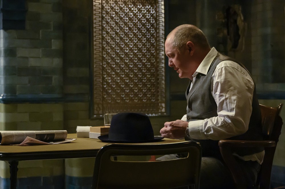 Raymond Reddington (James Spader) assis à une table manipule quelque chose avec ses mains