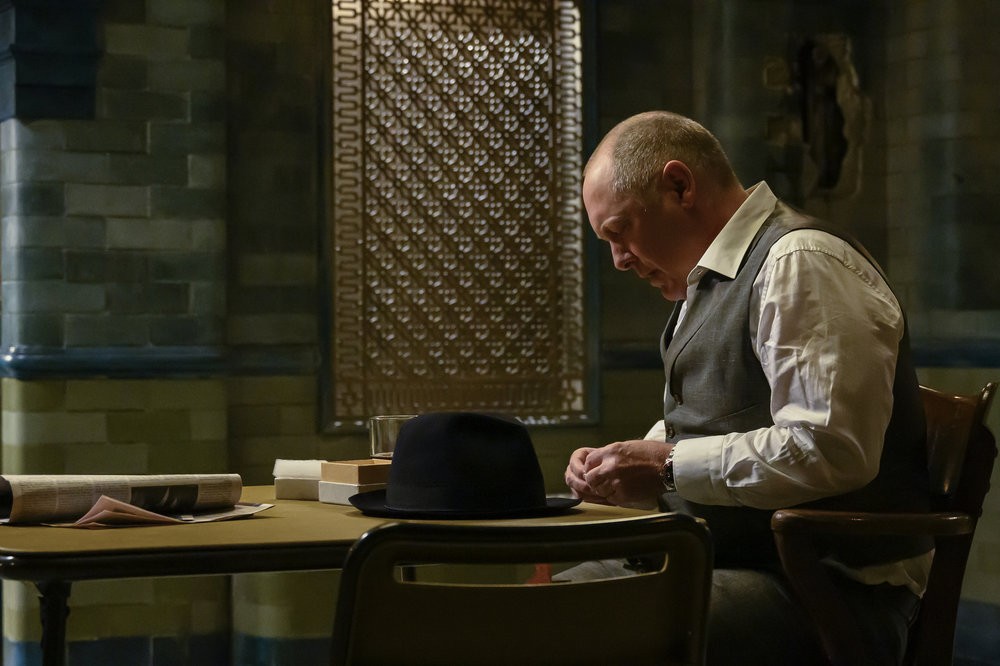 Reddington est assis à une table encombrée de divers objets dont son inséparable chapeau